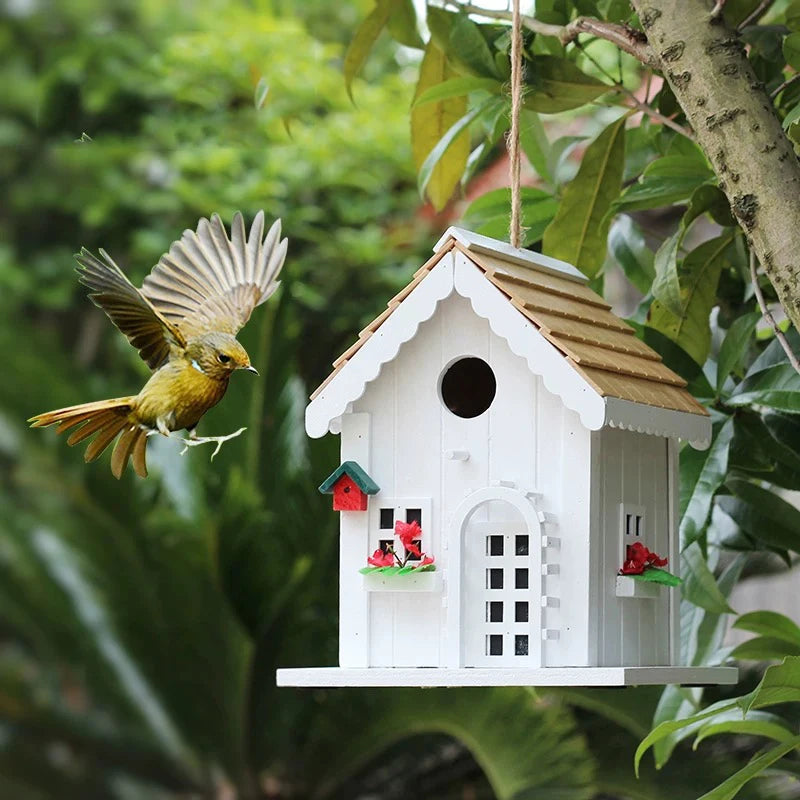 "Natural Wood House" wooden birdhouse hanging from tree with yellow bird in flight approaching entrance, jute rope hanger, red flower window boxes, lush green garden background