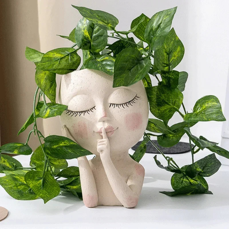 "Hush Muse" sculptural face resin planter in ivory cream displayed with lush cascading golden pothos plant as hair, on white desk with neutral background