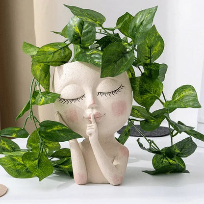"Hush Muse" sculptural face resin planter in ivory cream displayed with lush cascading golden pothos plant as hair, on white desk with neutral background