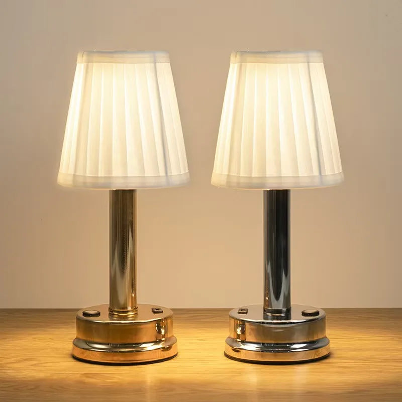  Two silver rechargeable touch table lamps with pleated fabric shades glowing side by side on a wooden surface, showing both Gold and Silver variants together