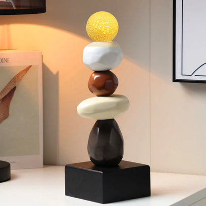 Abstract ceramic stacked sculpture with a yellow upper sphere, white and brown rounded forms, and a cream oval element on a black square base, displayed on a console in a softly lit modern interior.