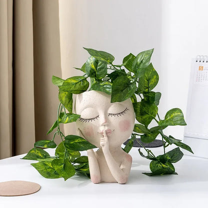 "Hush Muse" sculptural face resin planter in ivory cream with closed eyes and shh gesture, displayed with cascading golden pothos plant on white desk