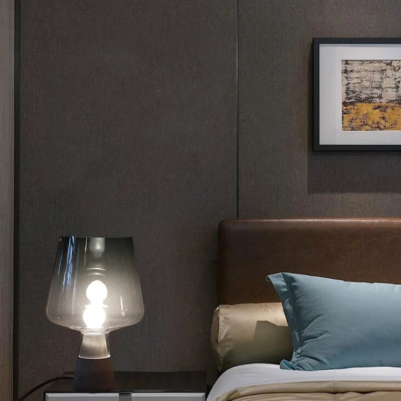 Illuminated smoked glass bedside table lamp with a dark textured base, styled on a nightstand beside a bed in a cosy modern bedroom.