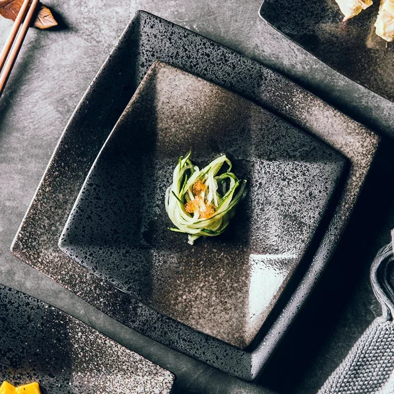 "Black Pearl" ceramic square dinner plate — top view lifestyle shot styled on a dark stone surface with a delicate Japanese appetiser at centre, chopsticks and linen napkin alongside, showing reactive glaze in deep black and silver tones — lifestyle shot