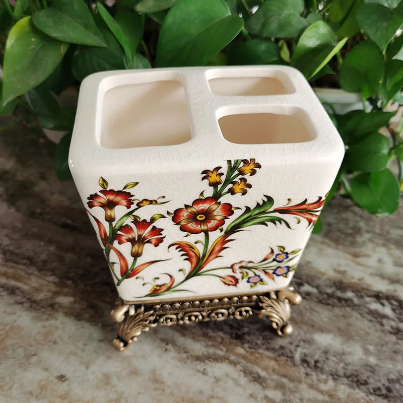 : White ceramic toothbrush holder with vibrant crimson and yellow Persian floral pattern on ornate bronze metal base, four slots, natural green leaf background