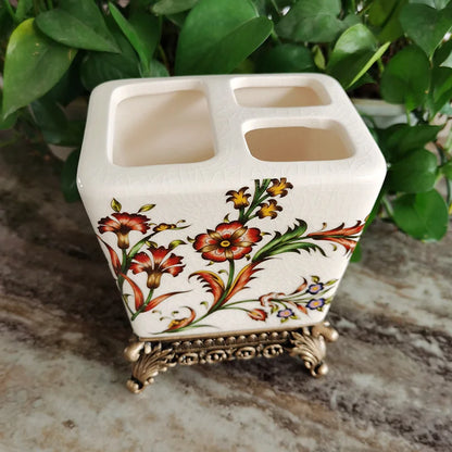 : White ceramic toothbrush holder with vibrant crimson and yellow Persian floral pattern on ornate bronze metal base, four slots, natural green leaf background