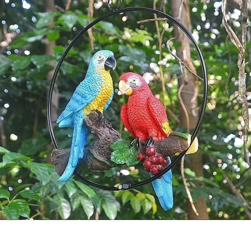 Perch Duo" resin parrot hanging ring ornament in Scarlet & Azure — hand-painted blue and red macaws perched on a driftwood branch with berry detail, garden background