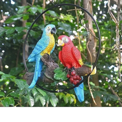 Perch Duo" resin parrot hanging ring ornament in Scarlet & Azure — hand-painted blue and red macaws perched on a driftwood branch with berry detail, garden background