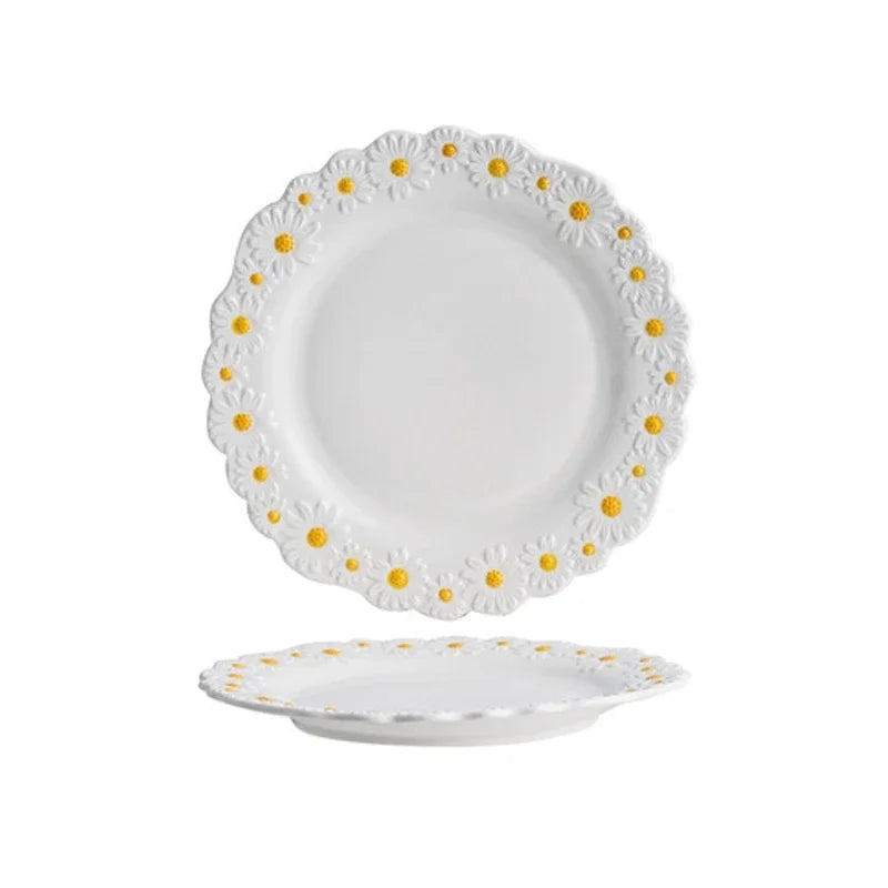 "Daisy Bloom" ceramic small plate — top and profile view on white background showing raised daisy relief border with white petals and golden yellow centres, soft white glaze and scalloped edge — product shot