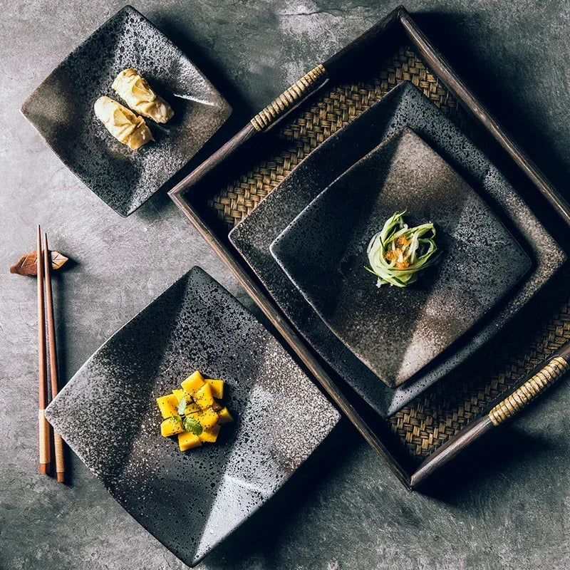 "Black Pearl" ceramic square dinner plate — lifestyle flat lay showing three plates in different sizes styled on a dark stone surface with Japanese appetisers, wooden chopsticks and woven rattan tray, displaying reactive glaze in deep black and silver speckled finish — lifestyle shot
