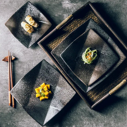 "Black Pearl" ceramic square dinner plate — lifestyle flat lay showing three plates in different sizes styled on a dark stone surface with Japanese appetisers, wooden chopsticks and woven rattan tray, displaying reactive glaze in deep black and silver speckled finish — lifestyle shot