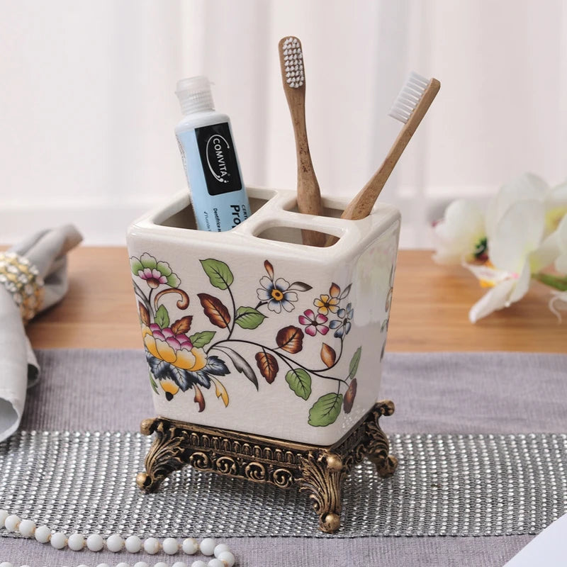 White ceramic toothbrush holder with hand-painted peony and botanical floral pattern in pink, yellow and green, ornate bronze metal base, two bamboo toothbrushes and toothpaste displayed, lifestyle background