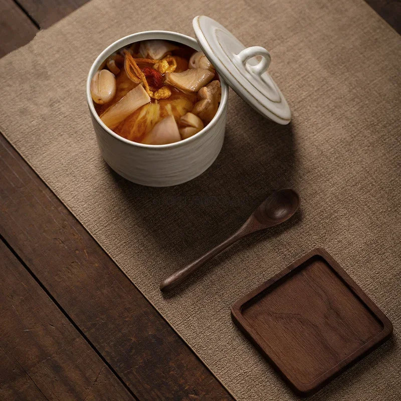 Ceramic lidded bowl filled with rich mushroom and vegetable broth, lid tilted to the side, styled on a linen table runner with a dark wooden spoon and small walnut tray, warm japandi setting