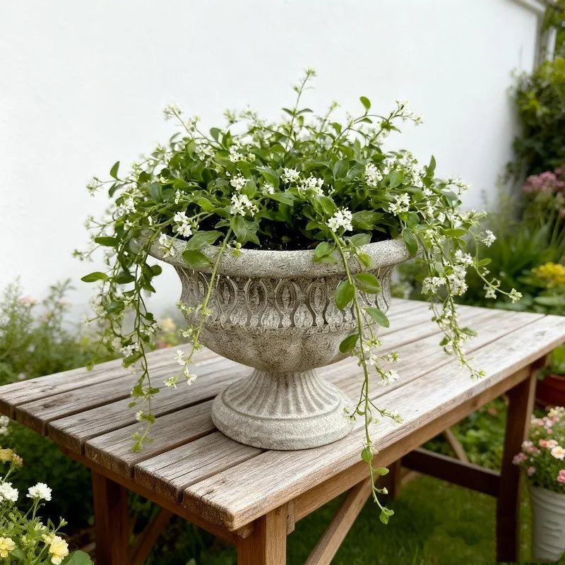 Roman Urn" ornate stone-texture garden urn planter styled with trailing white flowering plant, Stone Grey finish, placed on a rustic wooden garden table with lush garden background