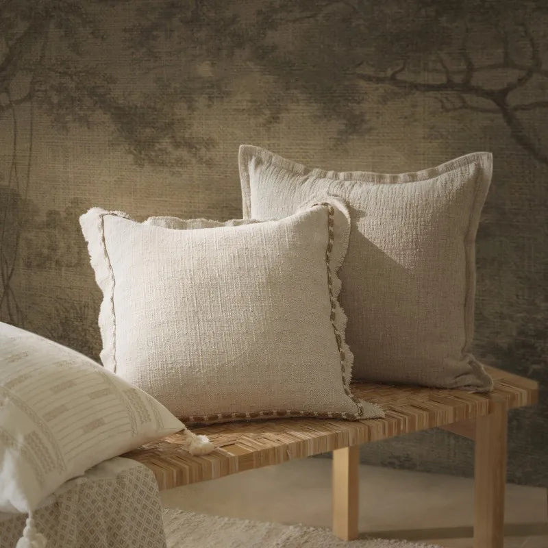 Two natural linen cushion covers with fringe edges layered on a woven rattan bench, styled against a vintage botanical mural wall — wabi-sabi and Japandi inspired home decor setting.