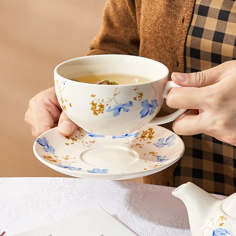 Hands holding a white porcelain teacup and saucer with blue chicory flowers and gold botanical details, with brewed tea inside, on a white table setting