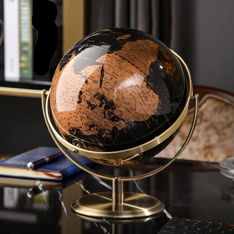 Decorative world globe with black and gold vintage map on gold-plated stand, styled on a dark glass desk with notebook and pen in a moody home office setting