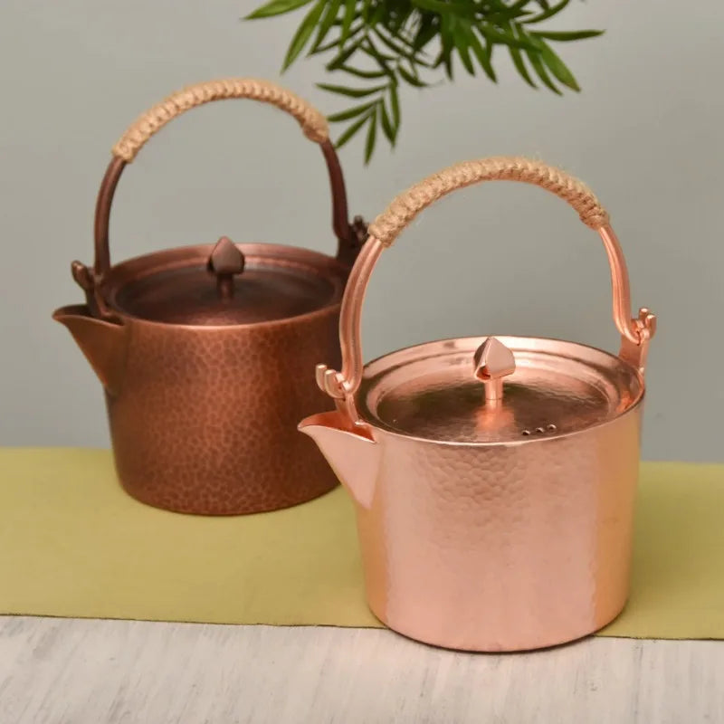 Copper Ritual handmade pure copper teapot kettle both variants side by side on a linen table runner — Rose Gold in front and Antique Copper behind — styled with a bamboo plant, showing the contrast between the two finishes