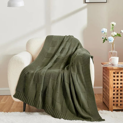 Chunky patchwork knit throw blanket in Forest Olive draped over a white bouclé armchair in a warm Scandinavian-inspired living room