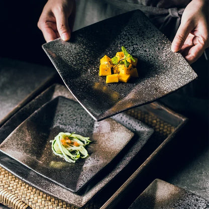 "Black Pearl" ceramic square dinner plate — lifestyle shot with chef's hands presenting a plate with mango and mint garnish over a dark surface, multiple plates stacked below with Japanese appetisers, showing reactive glaze in deep black and silver speckled finish — lifestyle shot

