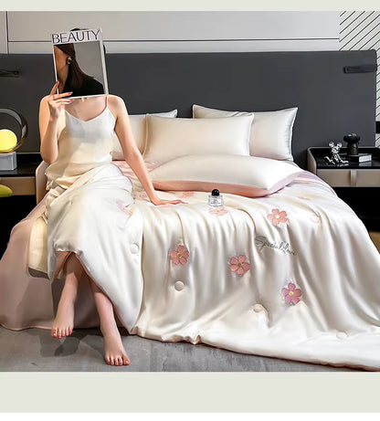 "Ice Bloom cooling summer quilt in Blossom Pink — woman sitting on bed with ivory white quilt draped over, embroidered pink daisy motifs and 'Special Love' script detail, blush pink reverse, elegant summer bedroom styling"