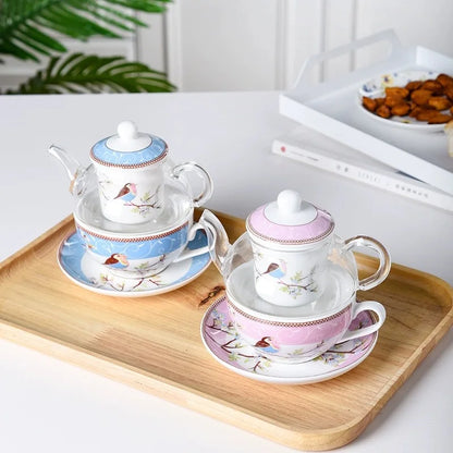 Blue and pink "Happy Bird" tea for one sets on a wooden tray, styled on a white surface — each with a glass teapot and porcelain cup and saucer, bird and blossom pattern, with tropical plant and snack bowl in the background.
