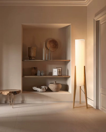 "Tripod & Cylinder" oak floor lamp casting warm light beside a built-in shelf with ceramic decorative objects in a neutral minimalist room