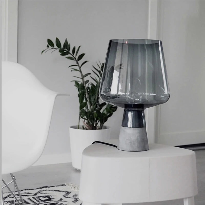 Smoked grey glass table lamp with a textured grey base styled on a white side table beside a potted plant in a modern interior.