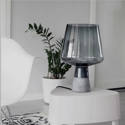 Smoked grey glass table lamp with a textured grey base styled on a white side table beside a potted plant in a modern interior.