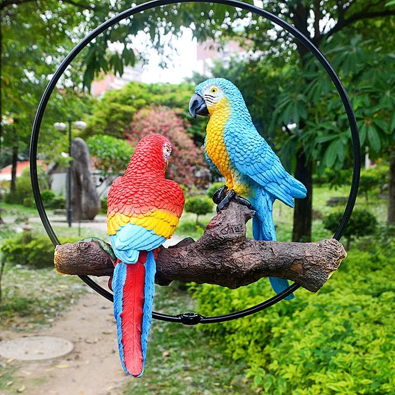 "Perch Duo" resin parrot hanging ring ornament in Scarlet & Azure — garden path setting showing full scale, red and blue macaws on log branch with lush green park background