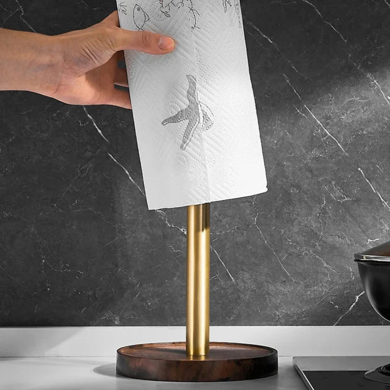Hand pulling a sheet of kitchen paper from the Walnut & Gold Paper Towel Holder on a white countertop, dark marble wall background, with a cooking pot to the side