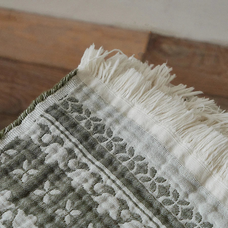 Close-up of white cotton fringe trim and jacquard floral weave detail on the Sage Bloom cotton throw blanket laid flat on a wooden surface