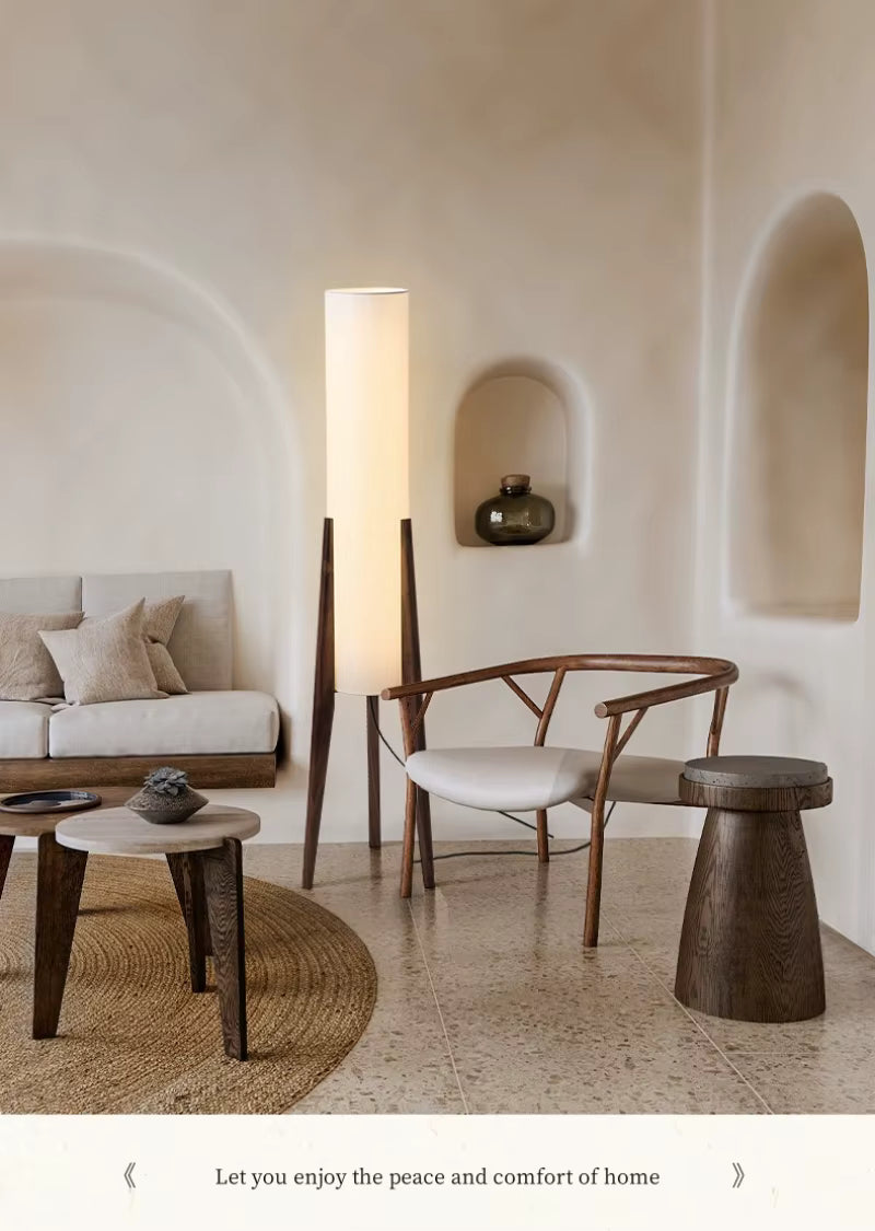 "Tripod & Cylinder" walnut floor lamp glowing warmly beside a wooden armchair and linen sofa in a serene wabi-sabi living room