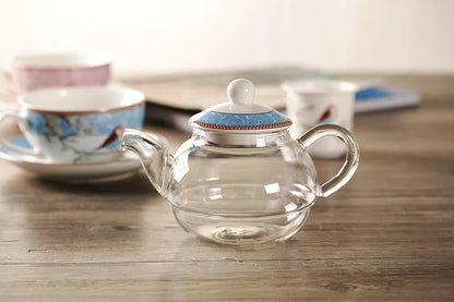 Clear glass teapot with blue porcelain lid from the "Happy Bird" tea for one set, on a wooden surface — blue cup and saucer softly blurred in the background.