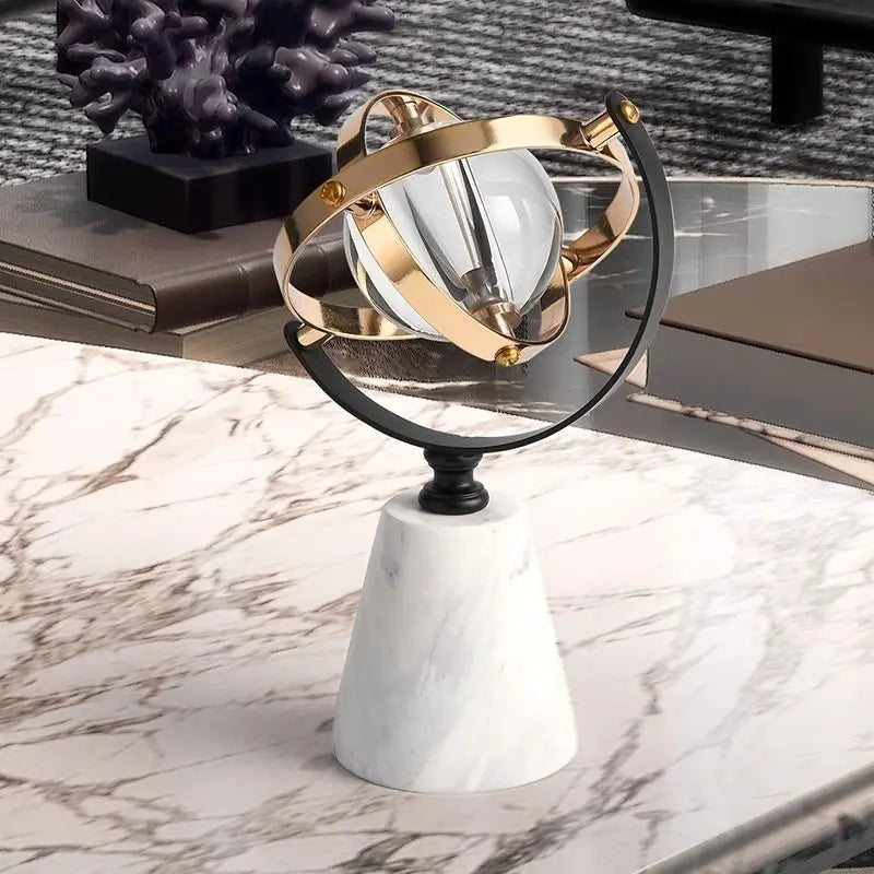 Decorative marble globe with a clear glass sphere and brass and black rings, on a marble coffee table beside books and a dark coral sculpture
