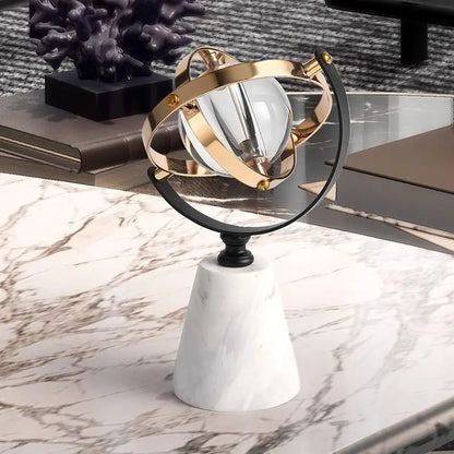 Decorative marble globe with a clear glass sphere and brass and black rings, on a marble coffee table beside books and a dark coral sculpture