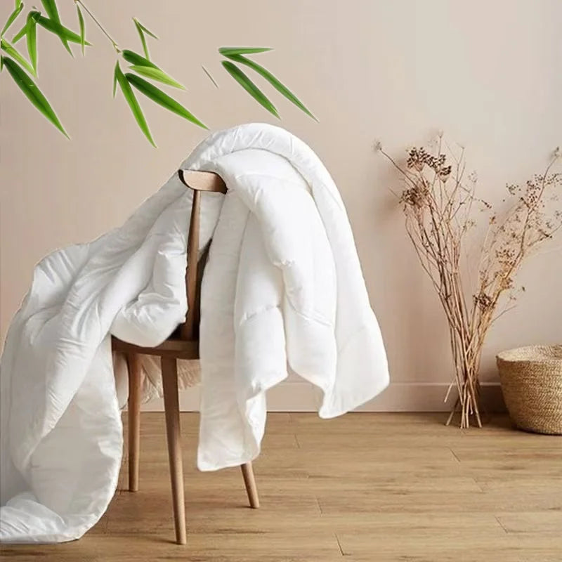  Ivory White 100% bamboo fibre cooling quilt draped over a wooden chair in a warm minimalist interior, with bamboo leaves overhead and dried botanicals beside it, evoking natural calm and effortless summer comfort
