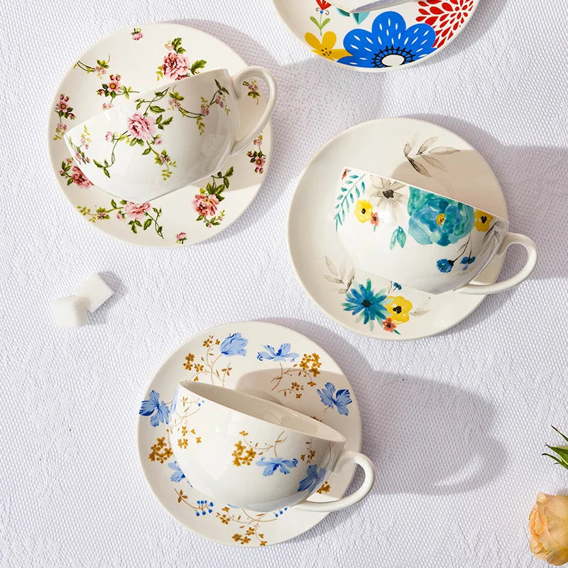 Tre floral porcelain tea cups and saucers displayed from above on a white linen surface, showing four different botanical designs including pink roses, blue chicory, watercolour blooms and folk flowers