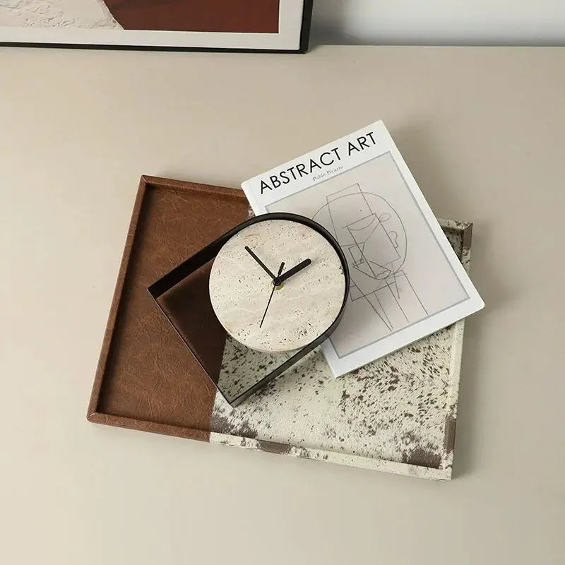 Black arched travertine table clock photographed from above, lying on an art book and a leather tray in a neutral styled setting