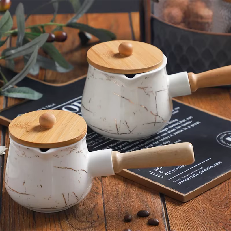  Two white marble ceramic milk jugs with bamboo lids and wooden handles displayed side by side on a dark slate board over a rustic wooden table, olive branch and coffee beans in the background — Small and Medium variants of the Marble Pour Ceramic Milk Jug by CL House