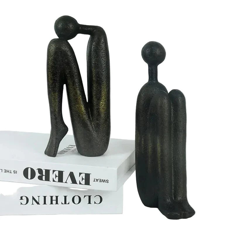 Pair of dark resin abstract human sculptures displayed as bookends on white books, with one figure standing and the other leaning forward.