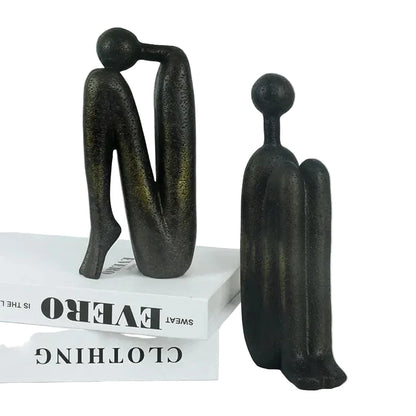 Pair of dark resin abstract human sculptures displayed as bookends on white books, with one figure standing and the other leaning forward.
