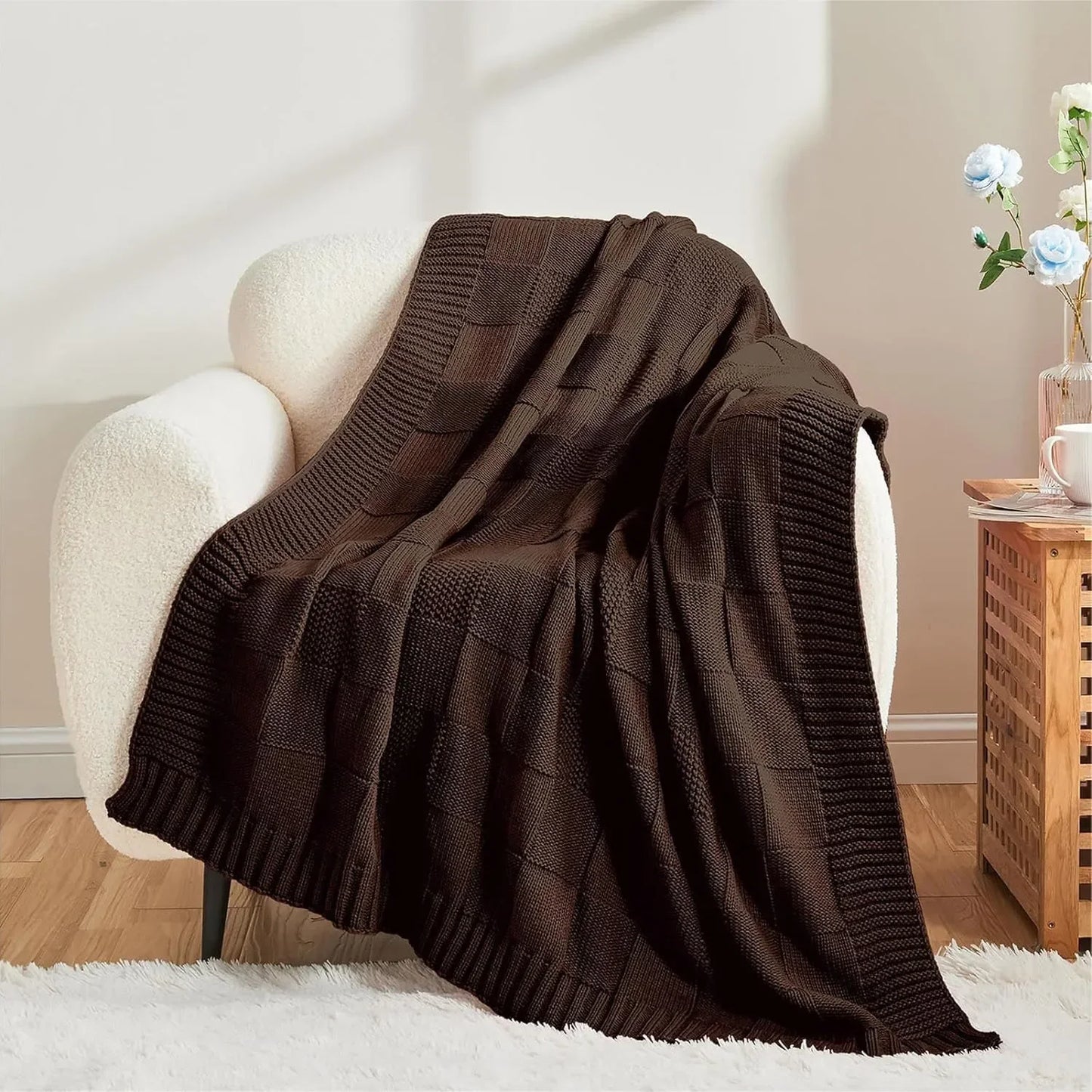 Chunky patchwork knit throw blanket in Dark Chocolate draped over a white bouclé armchair in a warm neutral living room with wooden side table
