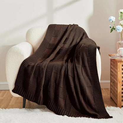 Chunky patchwork knit throw blanket in Dark Chocolate draped over a white bouclé armchair in a warm neutral living room with wooden side table