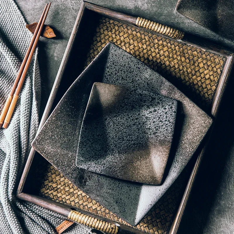 "Black Pearl" ceramic square dinner plate — top view lifestyle shot showing two plates nested on a woven rattan tray with wooden chopsticks and linen textile on a dark stone surface, displaying reactive glaze in deep black and silver speckled finish — lifestyle shot