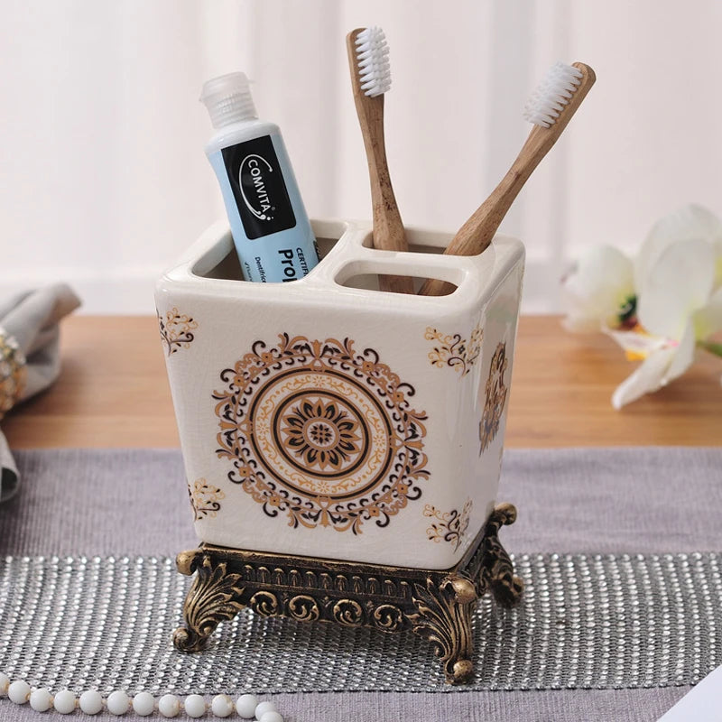 White ceramic toothbrush holder with golden medallion and arabesque pattern, ornate bronze metal base, two bamboo toothbrushes and toothpaste displayed, lifestyle background
