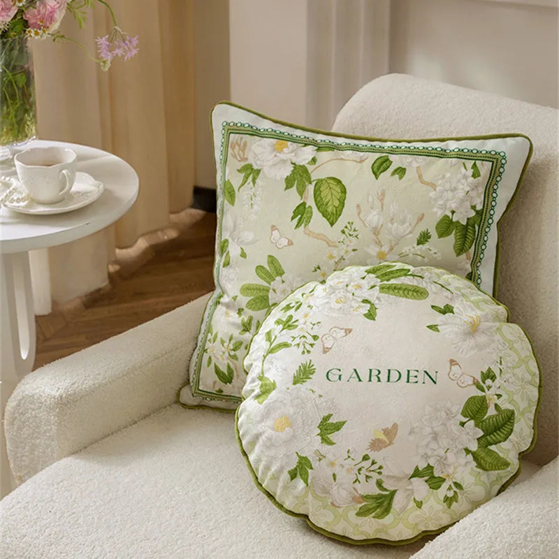  "Garden Whisper" cushion covers styled on a white bouclé armchair — Jasmine Garden Square and Round designs with white jasmine flowers and green foliage, scalloped round cushion with "GARDEN" script in foreground, pink flowers and tea cup in background