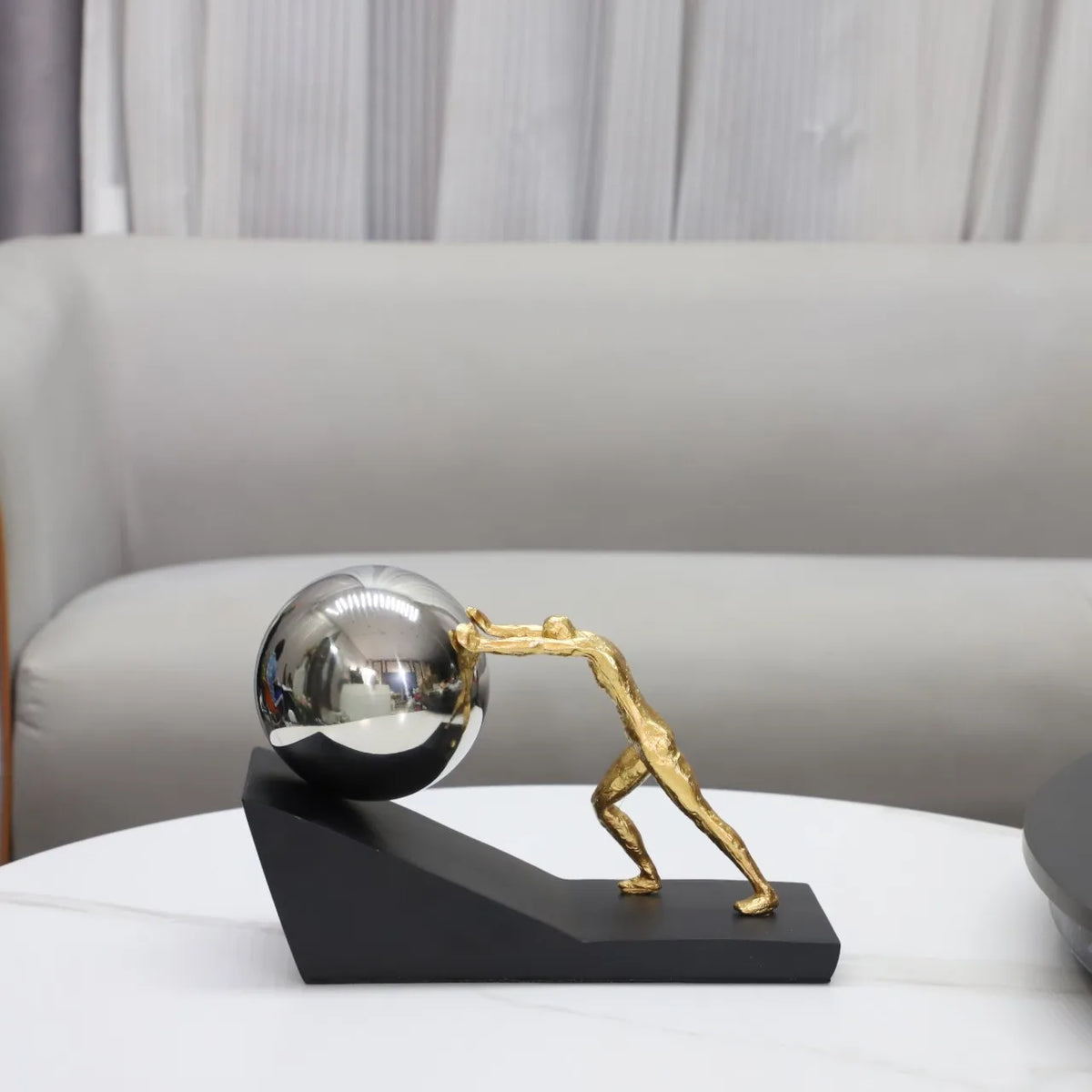 Silver Sphere version of The Ascent metal Sisyphus sculpture displayed on a white coffee table, with a cream sofa in the background.