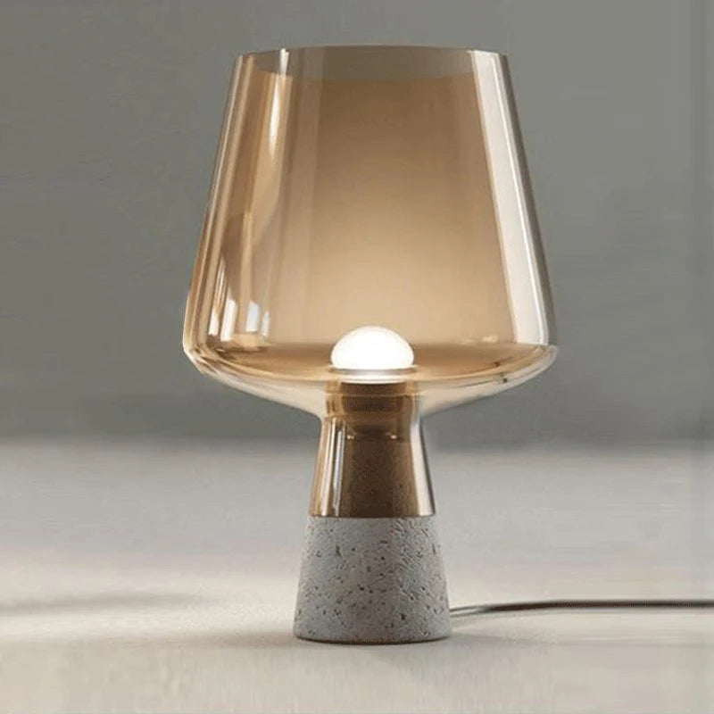 A modern table lamp with a wide amber glass shade and a warm glowing light inside, standing on a tapered grey speckled cement base on a light surface, with its power cord on the floor.