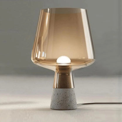 A modern table lamp with a wide amber glass shade and a warm glowing light inside, standing on a tapered grey speckled cement base on a light surface, with its power cord on the floor.